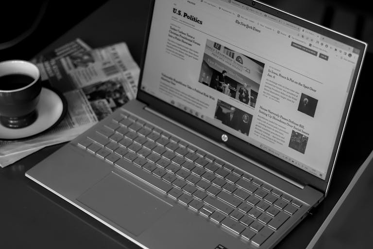 Black and white image of a laptop displaying news articles, accompanied by a cup of coffee and newspapers.